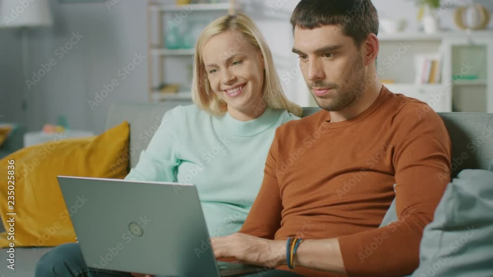 Cute Young Couple Sits on a Sofa at Home, They use Laptop Computer and Have Fun. Portrait of Beautiful Happy Young Couple in Love. Cute Young Couple Sits on a Sofa at Home, They use Laptop Computer and Have Fun. Portrait of Beautiful Happy Young Couple in Love.