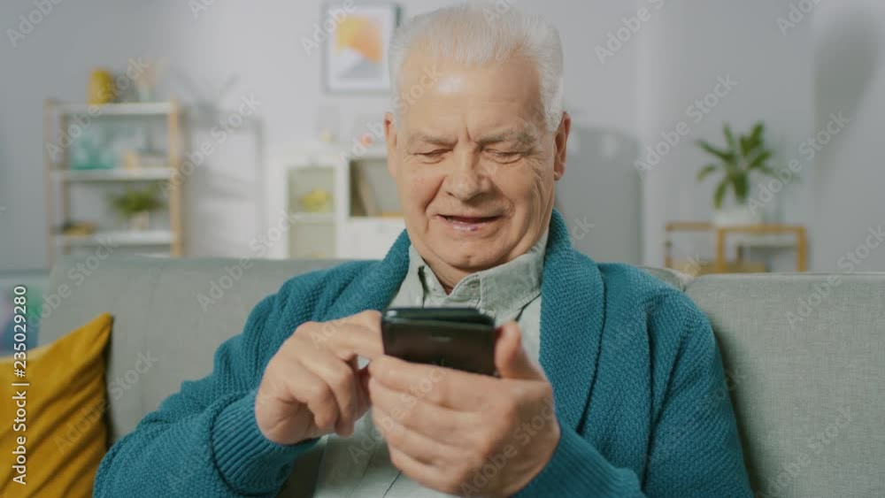 Portrait of Progressive Senior Man Sitting in His Living Room Easily Uses Smartphone, Does Touching and Swiping Gestures and Feels Very Comfortable with New Technologies. Portrait of Progressive Senior Man Sitting in His Living Room Easily Uses Smartphone, Does Touching and Swiping Gestures and Feels Very Comfortable with New Technologies.