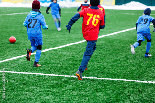 Fototapeta Naklejka Na Ścianę i Meble -  Young Active sport heathy boys in red and blue sportswear running and kicking a red ball on football field with artificial turf. soccer youth team plays football. activities for kids, training