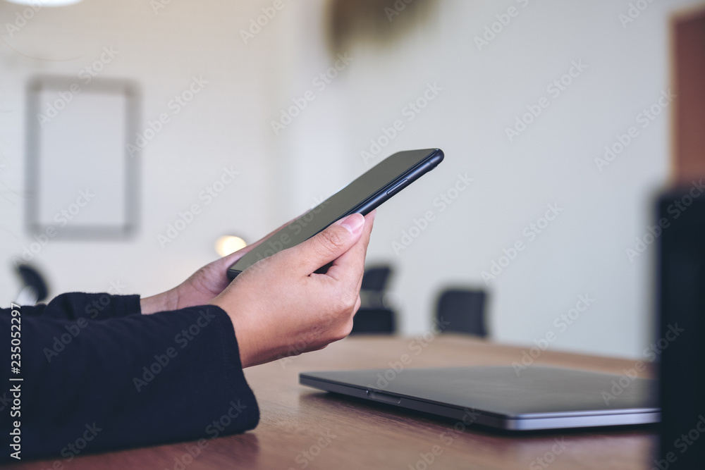 Fototapeta premium A woman holding and using black mobile phone with laptop on wooden table in office