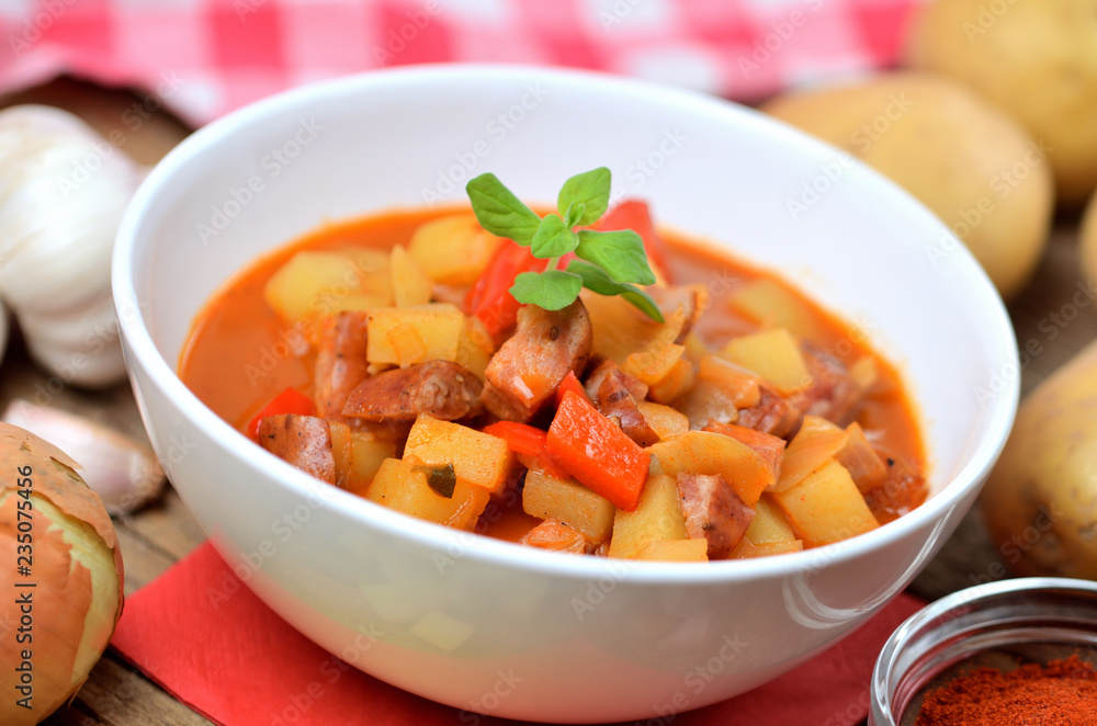 Potato and sausage goulash in bowl, pepper, onion and garlic on wooden table Potato and sausage goulash in bowl, pepper, onion and garlic on wooden table