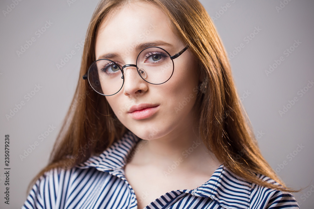 Beautiful closeup portrait of a young student girl wearing glasses on a gray background. Attractive redhead woman with natural beauty looks at the camera and smiles. Youth, clean skin