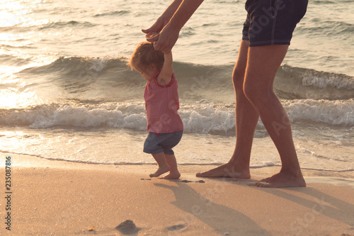 Father helping his baby with the first steps, teaching baby to walk concept, outdoor candid photo on the beach, spending a day at the beach, healthy family lifestyle.