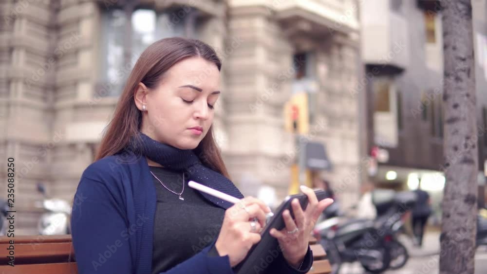 4k shot of a woman drawing on digital tablet with stylus pencil. Morern designer and paint freelance artist. Outdoors on the bench