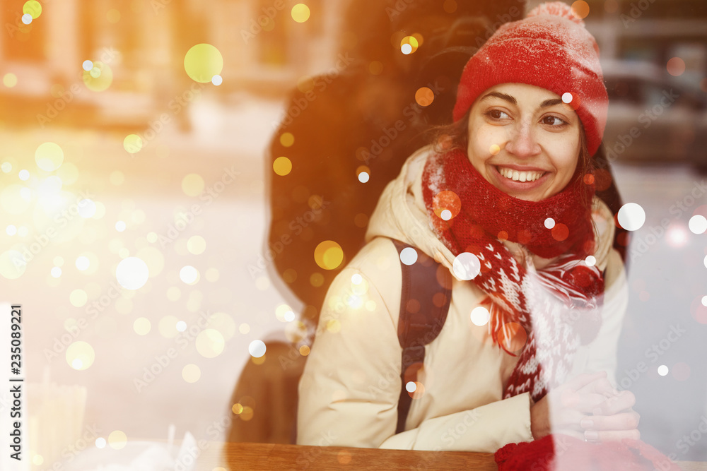 smiling happy woman in warm clothes, red knitted cap, scarf and mittens sitting in cafe near window and looking outside to the snowy street. view from outside, the window reflects snowy street smiling happy woman in warm clothes, red knitted cap, scarf and mittens sitting in cafe near window and looking outside to the snowy street. view from outside, the window reflects snowy street