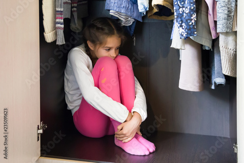Domestic violence and abused concept. Unhappy and scared little girl hiding in the wardrobe and sitting there down in the mouth 