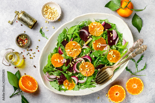 Gourmet salad of arugula, baked beetroot, tangerines and red onions with spices and pine nuts in a white dish. Olive oil and ingredients on a grey surface. Selective focus