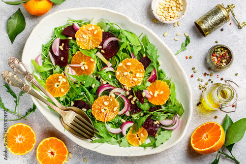 Delicatessen colorful salad of baked beets, arugula, tangerines and red onions with spices and pine nuts in a white dish. Olive oil and ingredients on a grey surface