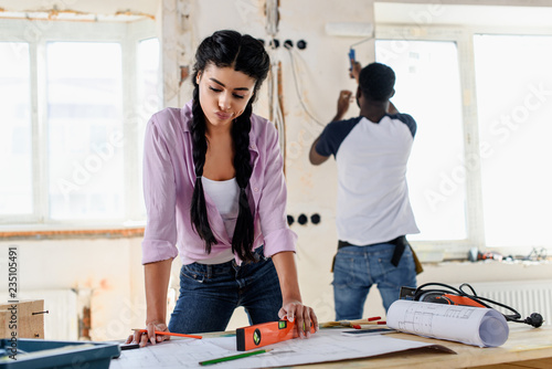 pensive young woman with spirit level looking at blueprint while her boyfriend working behind during renovation of home