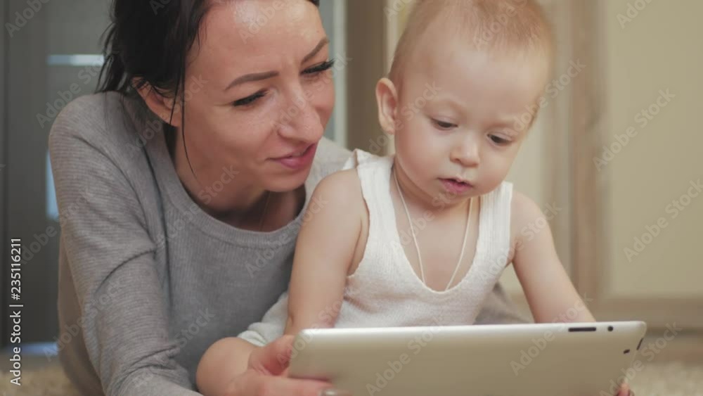 Young mother and baby boy play at tablet computer on a carpet at floor. Mother and son having fun with a digital tablet in home. Tablet computer and kids - mom showing son how to use touch interface. Young mother and baby boy play at tablet computer on a carpet at floor. Mother and son having fun with a digital tablet in home. Tablet computer and kids - mom showing son how to use touch interface.
