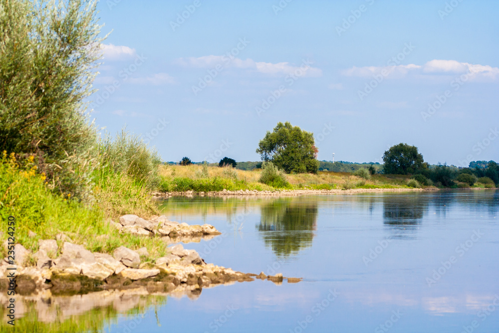 A beautiful river landscape. Flowing river that reflects the sky and plants on the shore. The shore is lined with stones. A beautiful river landscape. Flowing river that reflects the sky and plants on the shore. The shore is lined with stones.