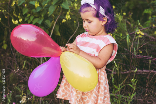 Child girl with purple hair in a dress holding multicolored balloons on a green natural background photo