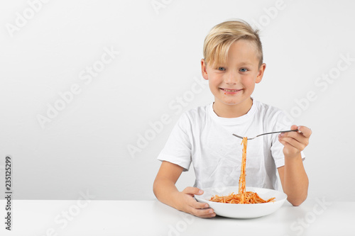 little blond boy in front of white background eats spaghetti with both hands and smiles