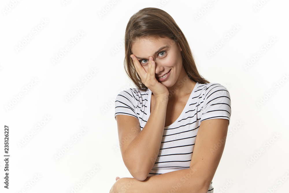 Charming girl in stylish sailor shirt flirting indoors, holding hand on her face and staring at camera, having playful or shy facial expression, smiling broadly, isolated against white wall background Charming girl in stylish sailor shirt flirting indoors, holding hand on her face and staring at camera, having playful or shy facial expression, smiling broadly, isolated against white wall background