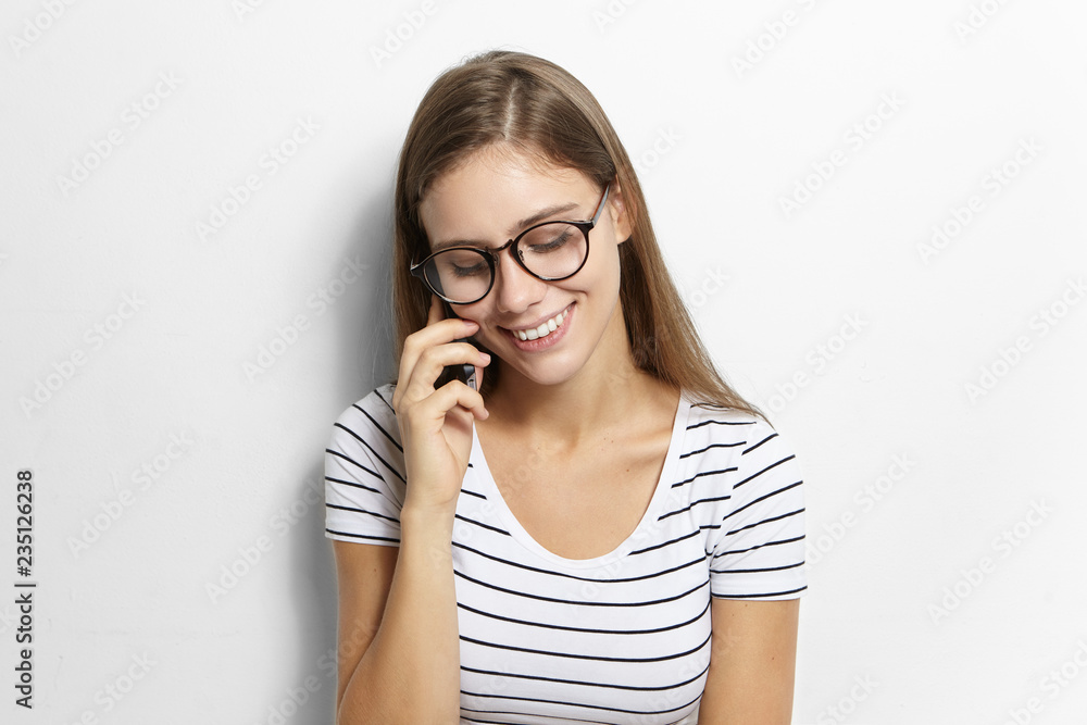Nice shy female teenager with long loose hair looking down and smiling broadly while talking on mobile phone to her boyfriend who asks her out on date. Youth, technology, gadgets and communication Nice shy female teenager with long loose hair looking down and smiling broadly while talking on mobile phone to her boyfriend who asks her out on date. Youth, technology, gadgets and communication