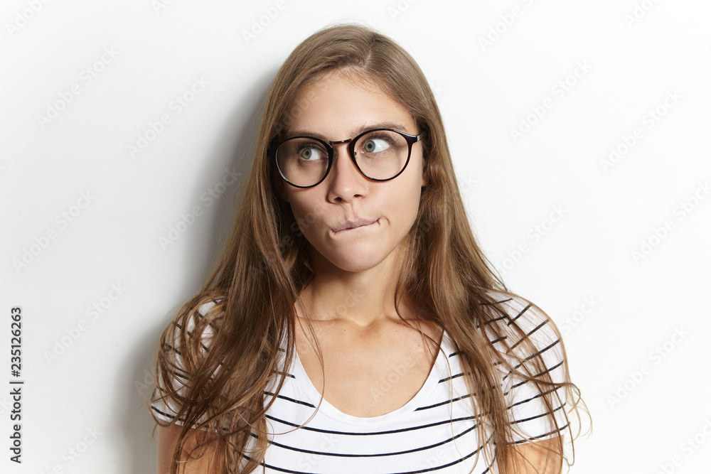Isolated shot of hesitant European girl in stylish eyeglasses pursing lips, having puzzled clueless facial expression, being deep in thoughts, thinking about exam, looking away. Human emotions