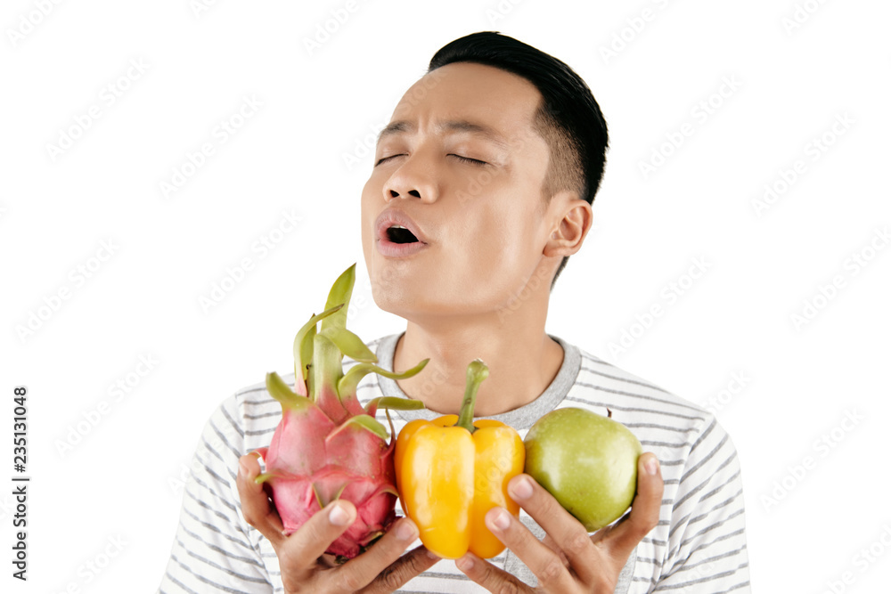 Waist-up portrait of young Asian man smelling fresh dragon fruit, apple and pepper with his eyes closed on white background Waist-up portrait of young Asian man smelling fresh dragon fruit, apple and pepper with his eyes closed on white background