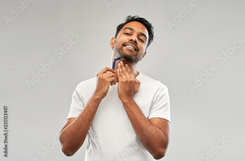 grooming and people concept - young indian man shaving beard with manual razor blade over grey background