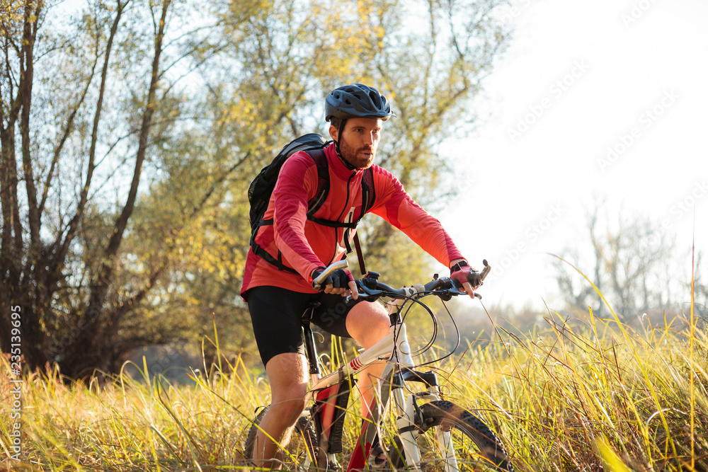 Determined young bearded man in red long sleeve cycling jersey riding mountain bike along a path through tall grass. Low angle front side view. Determined young bearded man in red long sleeve cycling jersey riding mountain bike along a path through tall grass. Low angle front side view.
