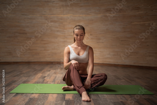 Peaceful young woman in comfortable clothes feeling relaxed while sitting on the yoga mat in empty room