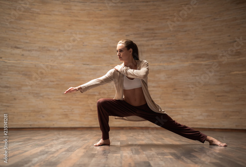 Fototapeta Naklejka Na Ścianę i Meble -  Tranquil slim young lady bending her knee and looking into the distance while putting one hand on the shoulder during the chi gong practice