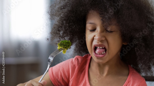 Funny African girl eating broccoli with huge disgust, healthy diet for children