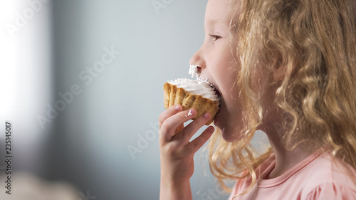 Quadro su tela Cute pale preschool girl eating creamy cake and smiling, candy bar at party