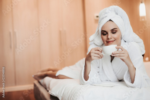 Concept of relaxation and harmony after bath. Full length portrait of young smiling lady in bathrobe lying on bed with cup of tea. Copy space on left