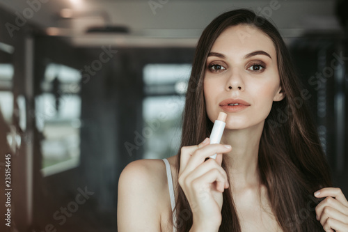 Portrait of serene young female with long hair and dark eyes applying make up in apartment