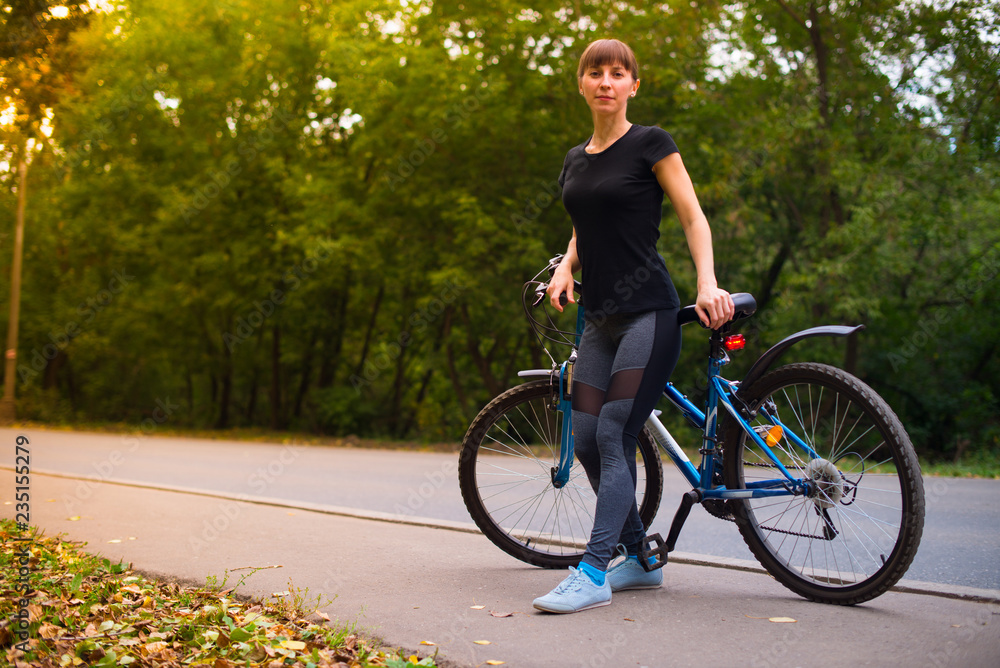 Young beautiful athletic girl in sportswear sitting on a blue bike frame. Trees on the background.