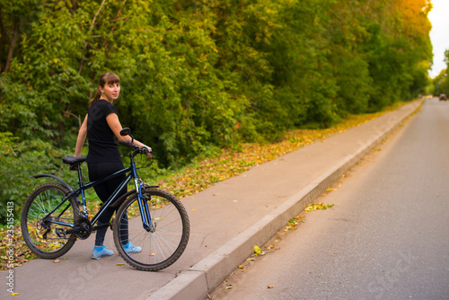 Young beautiful athletic girl in sportswear turned around sitting on the frame of a blue bicycle. Trees on the background.