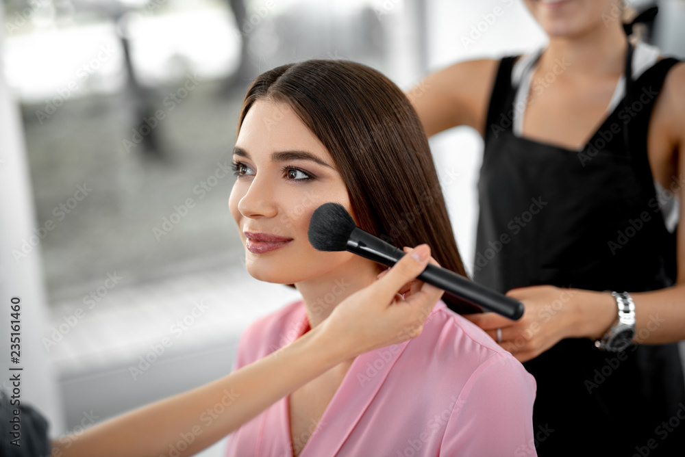 Make up. Young long haired woman sitting at the beauty salon and smiling while professional beautician putting rouge on her cheek
