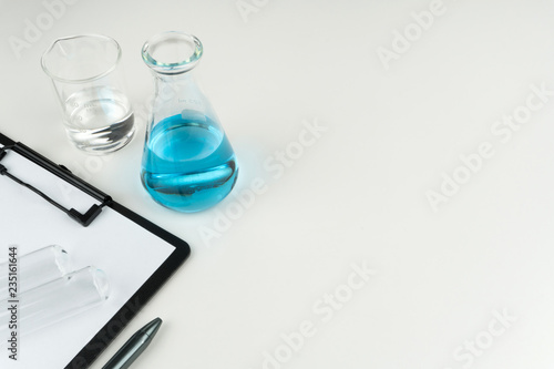 laboratory equipments. A blue liquid flask, clear liquid beaker, test tubes and a clipboard with pen on the white table