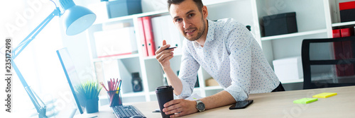 A young man is standing near a table in the office, holding a pencil and a glass of coffee. A young man works with documents and a computer. photo