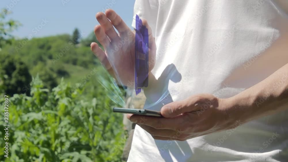 Man shows concept hologram Blockchain on his phone. Person in white t-shirt with future technology holographic screen and green nature background Man shows concept hologram Blockchain on his phone. Person in white t-shirt with future technology holographic screen and green nature background