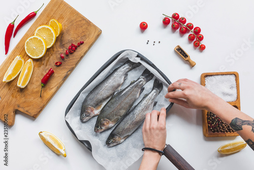 cropped image of woman putting uncooked fish in tray with baking paper on white table with ingredients