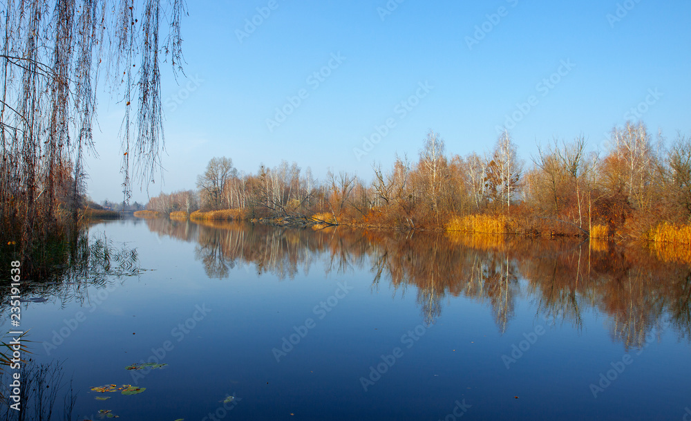 Beautiful autumn trees reflecting on the smooth water surface. Warm autumn day on the lake. River bank landscape.