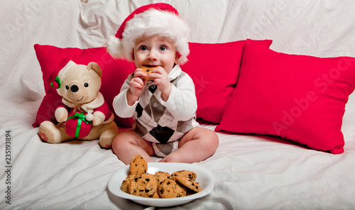A little kid in a red cap eats a cookies and milk. Christmas photography of a baby in a red cap. New Year holidays and Christmas