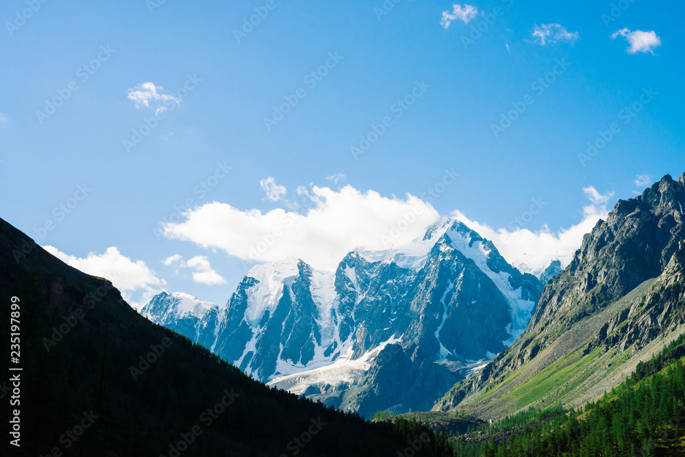 Amazing glacier under blue sky. Huge cloud on giant snowy mountains in sunlight. Rich vegetation and forest of highlands. Atmospheric minimalist mountain landscape of majestic nature in sunny day.