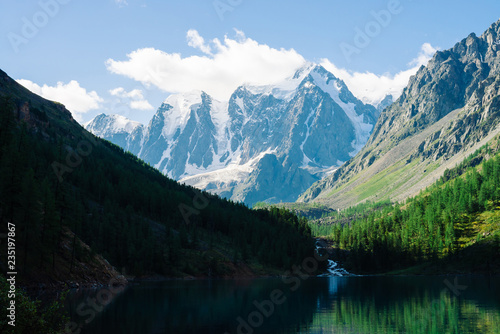 Amazing glacier under blue sky. Forest reflected on clean water of mountain lake. Huge cloud on giant wonderful snowy mountains. Rich vegetation of highlands. Atmospheric landscape of majestic nature.