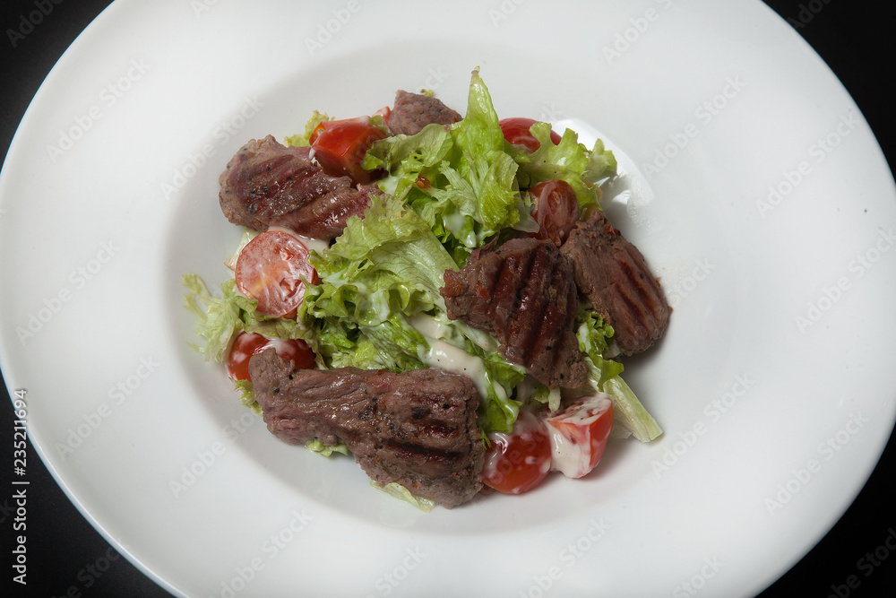 Veal salad, salad with large slices of veal and fresh vegetables on a plate on a black background.