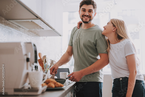 Good morning for two. Attractive smiling guy cooking food for his gorgeous girlfriend. She standing nearby and hugging him with love