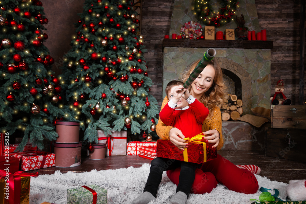 Young mother female woman playing with son child kid in red sweater at home near decorated Christmas tree and fireplace during winter holidays. Merry Christmas and Happy New Year concept,