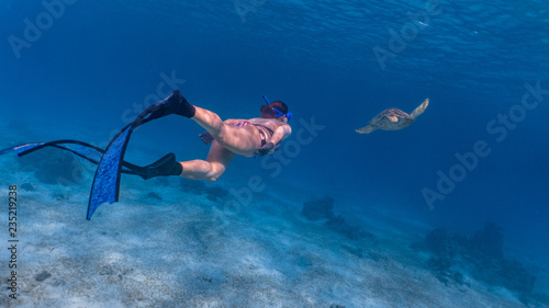 A lady free diver in sexy bikini chasing a green sea turtle in a sandy reef