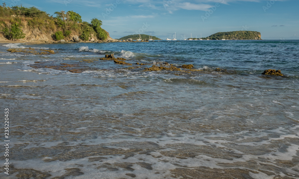Fototapeta premium View from the water of waves coming into shore with rocky headlands, sailboats, and islands along the horizon on a sunny day in the Caribbean.