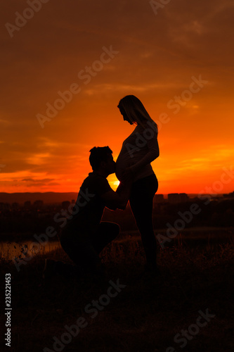 Silhouettes of husband kissing stomach of his pregnant wife while they enjoy spending time together outdoor.