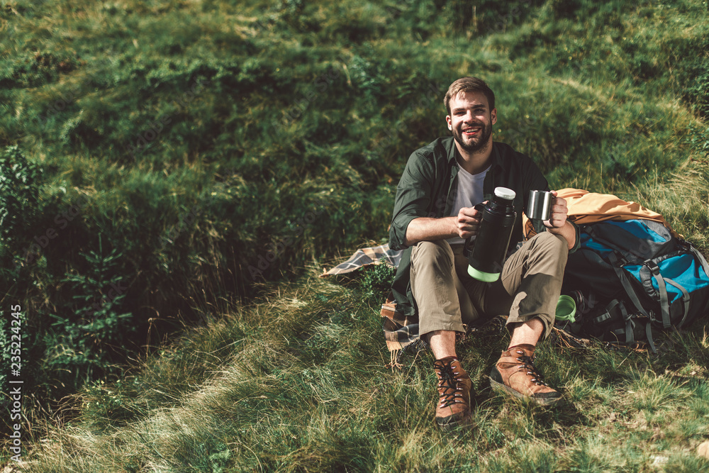 Take a break in journey and adventure. Full length portrait of young traveler male holding cup and thermos bottle while camping on green hill. Copy space on left