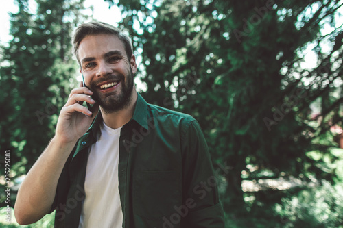 Communication during interesting travelling. Close up portrait of young cheerful man talking by mobile phone while standing on green trees background