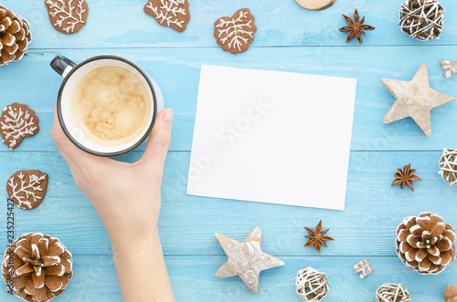 Christmas Mock up blank white paper sheet. Top view of woman's hands holds coffee. Pine cone,wood star on blue background.Christmas winter new year and xmas greeting card wish. Flat lay