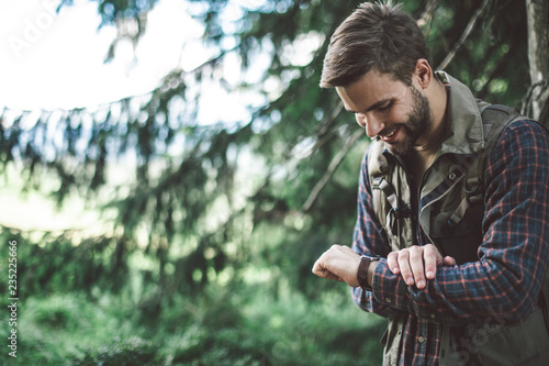 Being in time in journey and adventures. Side on portrait of smiling male traveler walking uphill in green forest and looking at his watch. Copy space on left